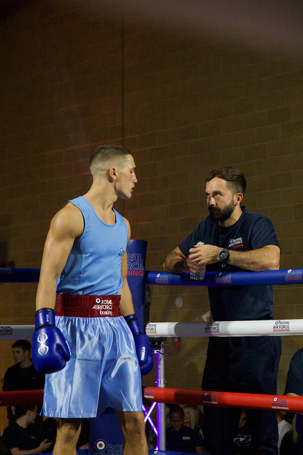 An RAF amateur boxer stands in an allotted ring corner, talking with a coach who is outside the ring.